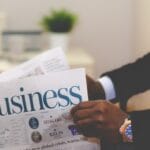 Person in a suit holding a newspaper with the headline Business; a potted plant is blurred in the background.