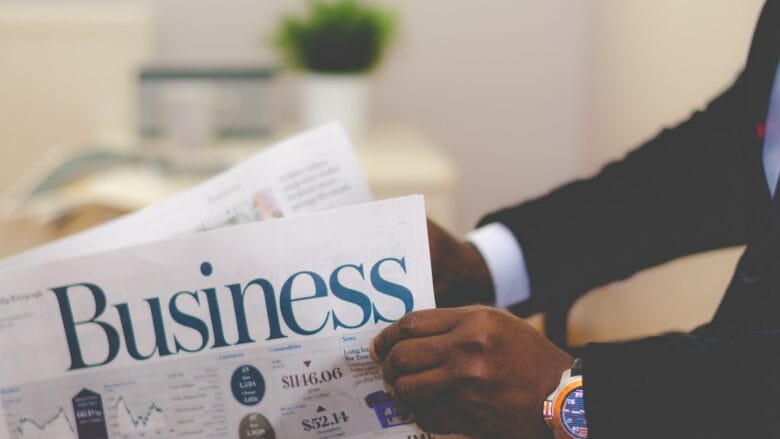 Person in a suit reading a newspaper with the headline Business; charts and financial news are visible. A potted plant is in the background.