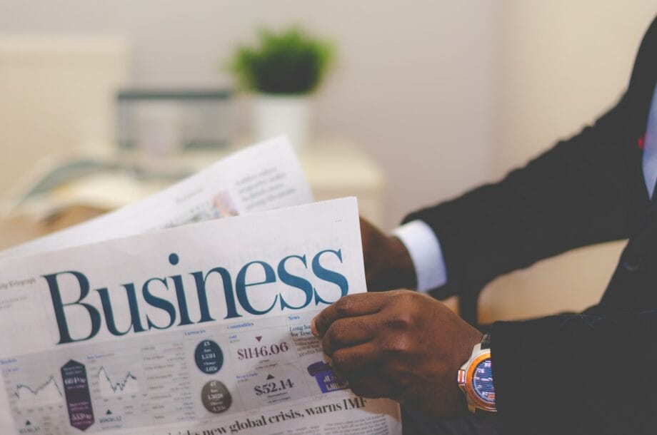Person in a suit reading a newspaper with the headline Business; charts and financial news are visible. A potted plant is in the background.