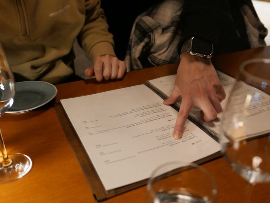 Two people sit at a table; one person points at a menu while the other looks on. Glasses and plates are on the table.