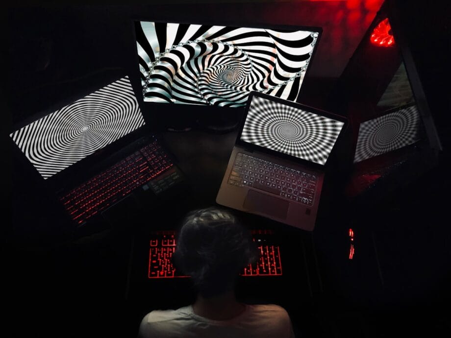 A person sits at a desk with four screens displaying black and white spiral patterns, illuminated by red keyboard lights.