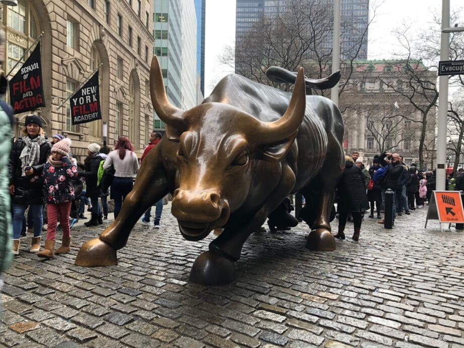 The Charging Bull statue stands on cobblestone with a crowd of people gathered around it in an urban setting.