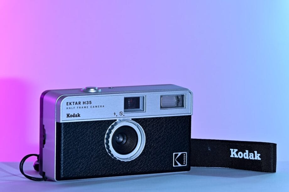 A Kodak Ektar H35 half frame camera with a Kodak-branded strap, placed on a flat surface against a pink and blue gradient background.