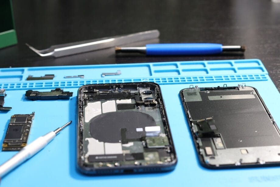 Disassembled smartphone with internal components, tools, and a blue work mat on a black surface.