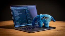A laptop with code on the screen sits on a table with a blue PHP elephant plush toy placed on the keyboard.