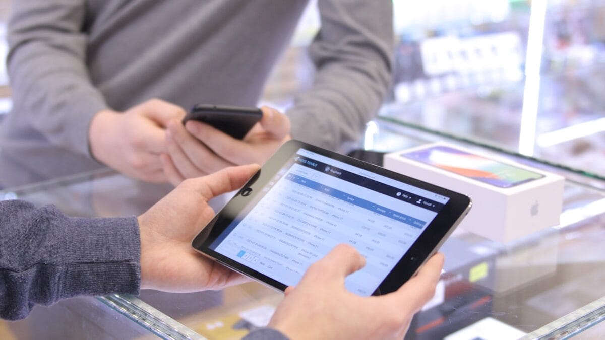 Two people using a smartphone and a tablet at a glass display counter, with a boxed device visible in the background.