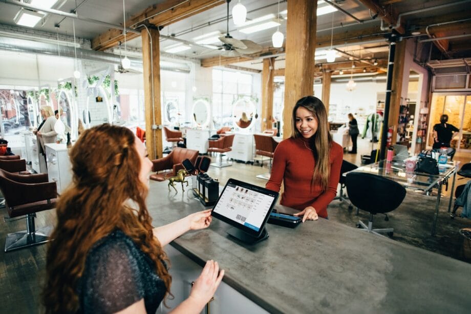 Two women interact at a salon reception desk, with one using a touchscreen computer and the other standing and smiling.