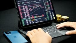 Person using a tablet displaying Binance cryptocurrency trading charts, with gold coins and a blue smartphone nearby on a black surface.