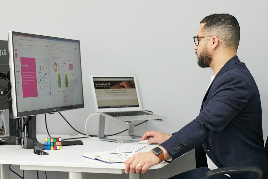 A man sits at a desk working on a computer and laptop, with documents, a notebook, and a Rubik’s Cube nearby.