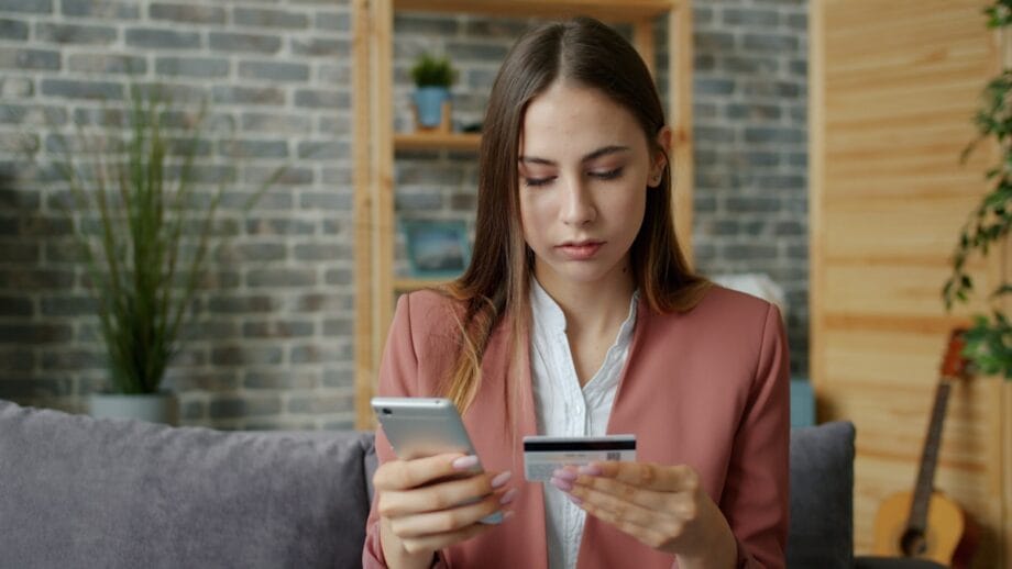 A woman sits on a couch holding a smartphone and credit card, appearing to make an online transaction.