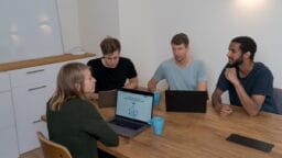 Four people sit around a table with laptops and mugs, engaged in a discussion in a meeting room.