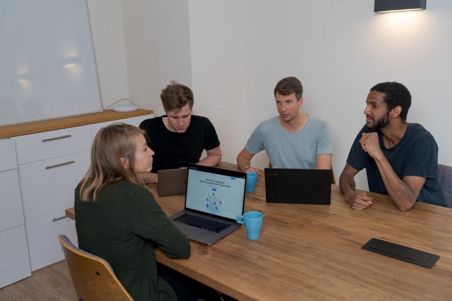Four people sit around a table with laptops and mugs, engaged in a discussion in a meeting room.