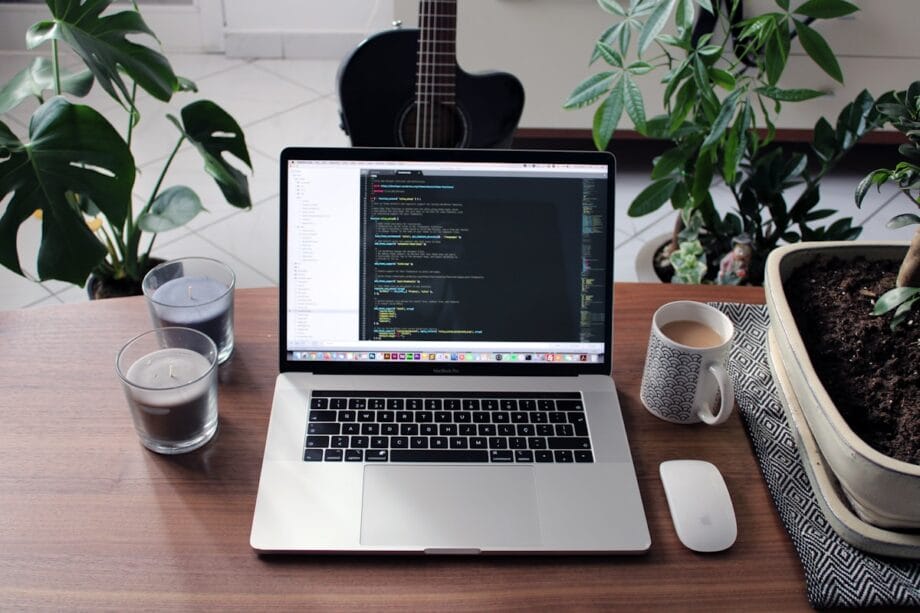 A laptop displaying code sits on a wooden table with candles, a mug, a computer mouse, houseplants, and a guitar in the background.