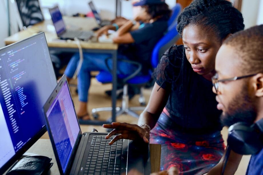 Two people work together at a desk, looking at code on a laptop screen; others work on computers in the background.