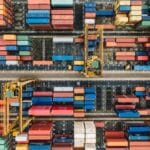 Aerial view of numerous colorful shipping containers organized in rows at a busy port with cranes for loading and unloading.