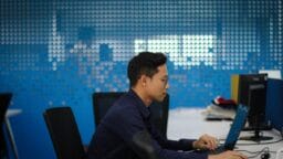 Man wearing glasses sits at a desk typing on a laptop in a modern office with blue walls and multiple computer stations.