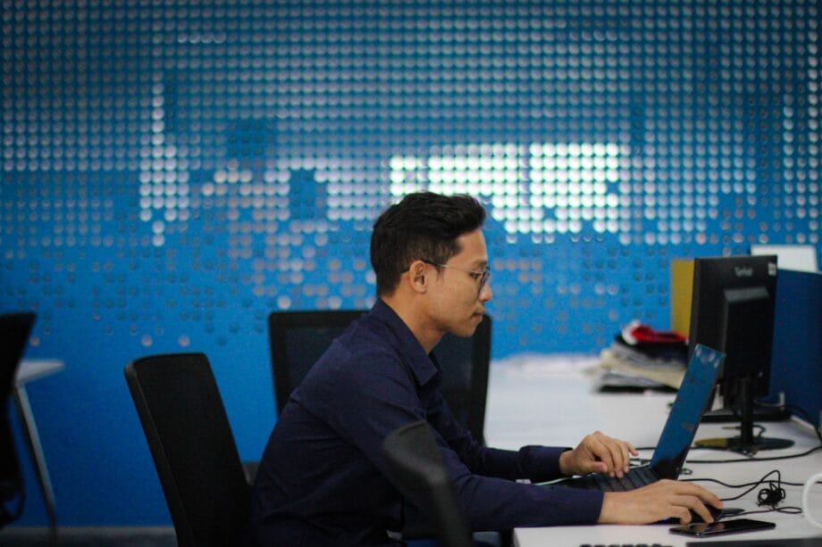Man wearing glasses sits at a desk typing on a laptop in a modern office with blue walls and multiple computer stations.