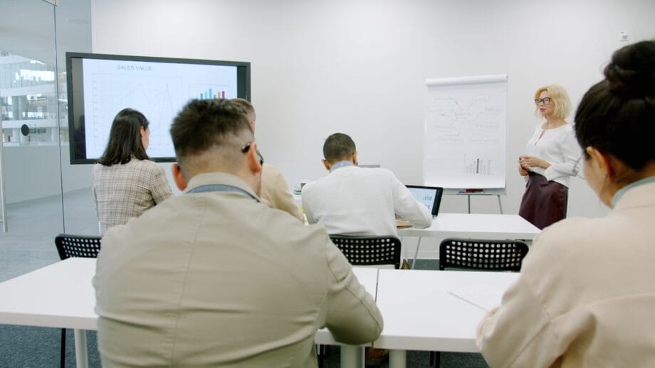 A woman presents sales data to a group of people seated at desks in a modern office meeting room.