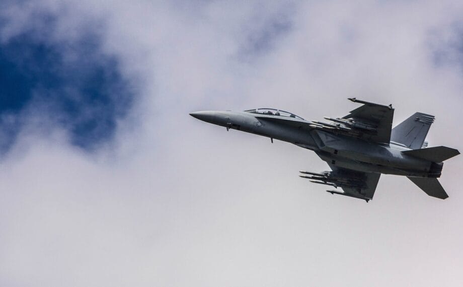 A military fighter jet with missiles attached flies through a partly cloudy sky.