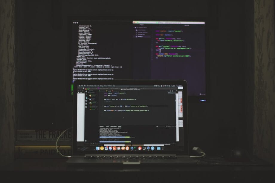 A laptop and a monitor display lines of code in a dimly lit room, with stacked books visible beside the laptop.