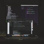 A laptop and a monitor display code editors with programming code in a dimly lit workspace.