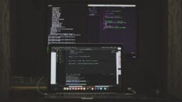 A laptop and a monitor display code editors with programming code in a dimly lit workspace.