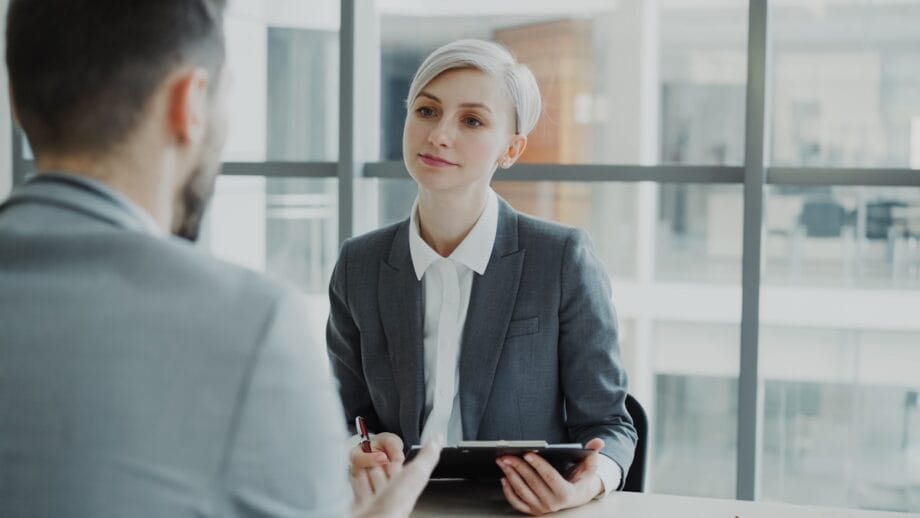 A woman in a gray suit holds a tablet and listens to a man during a business meeting in a modern office.