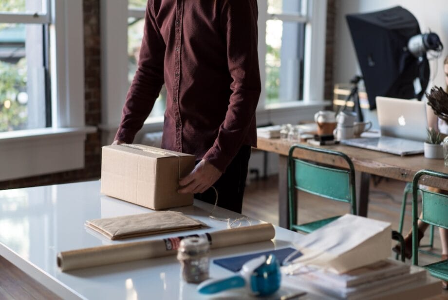 Person standing at a desk, packing a cardboard box in a workspace with office supplies, papers, and equipment visible.