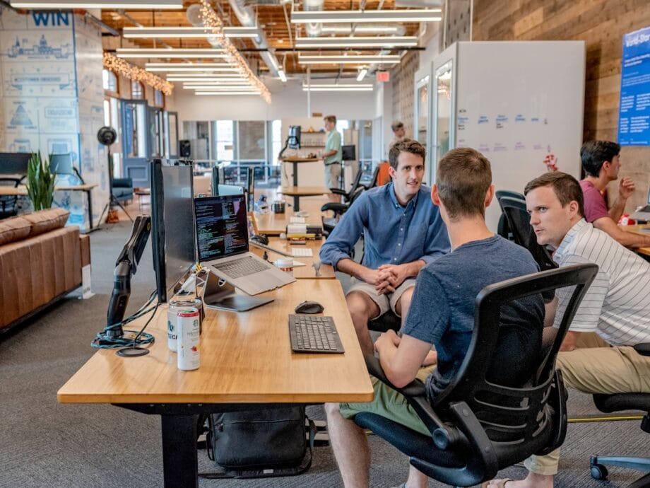 Three men have a discussion at a desk with computers in a modern, open-plan office with other people working in the background.