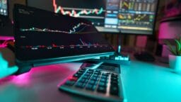 A desk with a calculator, computer monitor, and tablet displaying stock market charts and trading data.