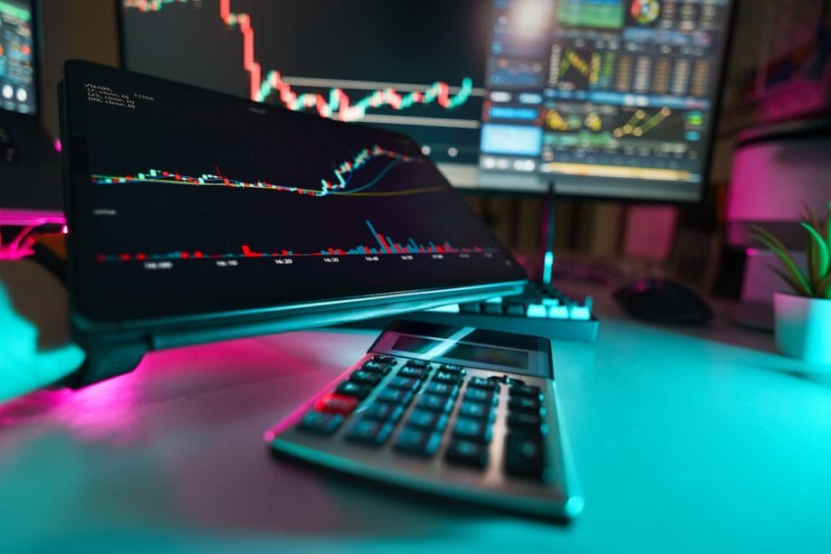 A desk with a calculator, computer monitor, and tablet displaying stock market charts and trading data.