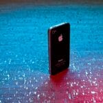 An older model black iPhone is standing upright on a textured blue and pink illuminated surface.