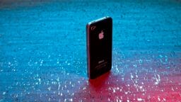 An older model black iPhone is standing upright on a textured blue and pink illuminated surface.