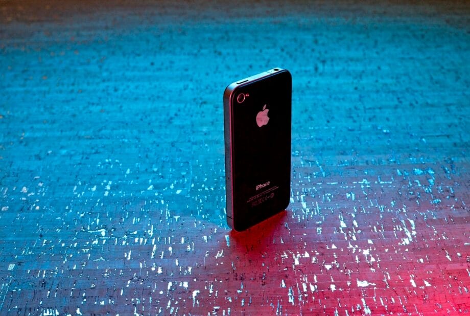 An older model black iPhone is standing upright on a textured blue and pink illuminated surface.