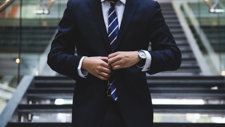 A person in a dark suit and striped tie buttons their jacket, standing in front of a modern staircase.