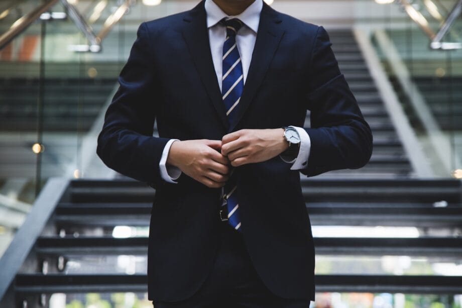 A person in a dark suit and striped tie buttons their jacket, standing in front of a modern staircase.