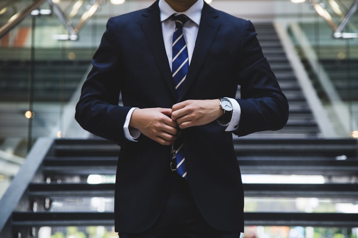 A person in a dark suit and striped tie buttons their jacket, standing in front of a modern staircase.