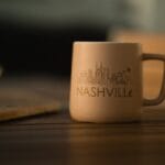 A ceramic mug with a simple Nashville city skyline illustration and the word Nashville printed on it sits on a wooden table.