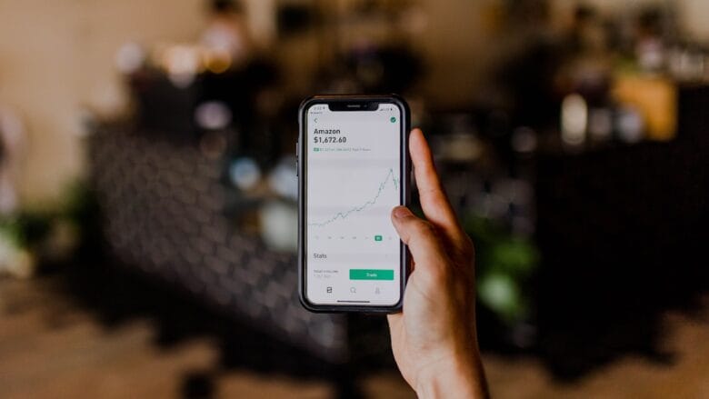 A hand holds a smartphone displaying a stock trading app with an upward trending Amazon stock chart on the screen.