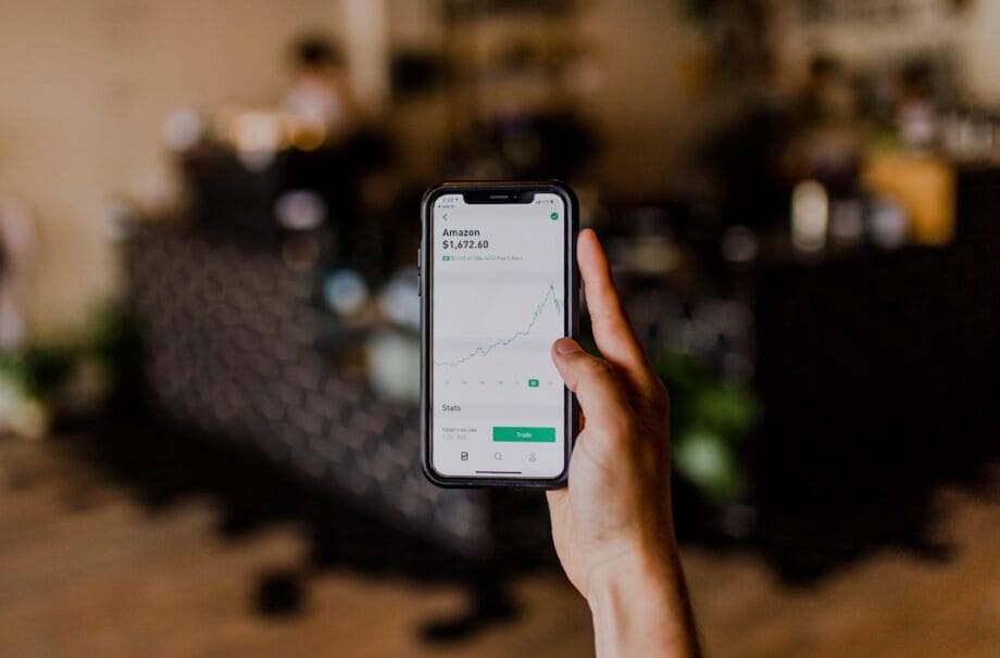 A hand holds a smartphone displaying a stock trading app with an upward trending Amazon stock chart on the screen.