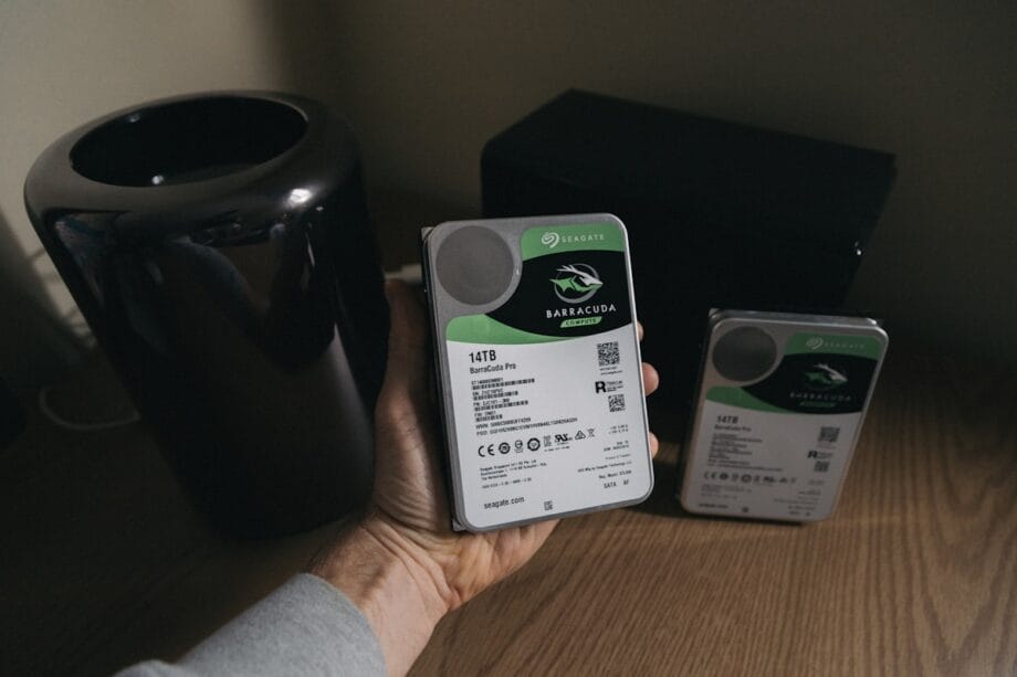A person holds a 14TB Seagate Barracuda hard drive; another similar hard drive rests on a wooden surface nearby.