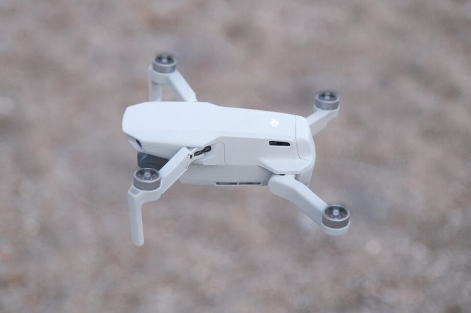 A small white quadcopter drone is flying above a blurred, gravelly surface.