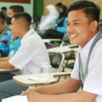 Male student in a white shirt smiles while sitting in a classroom with other students, some of whom are wearing uniforms.