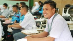 Male student in a white shirt smiles while sitting in a classroom with other students, some of whom are wearing uniforms.
