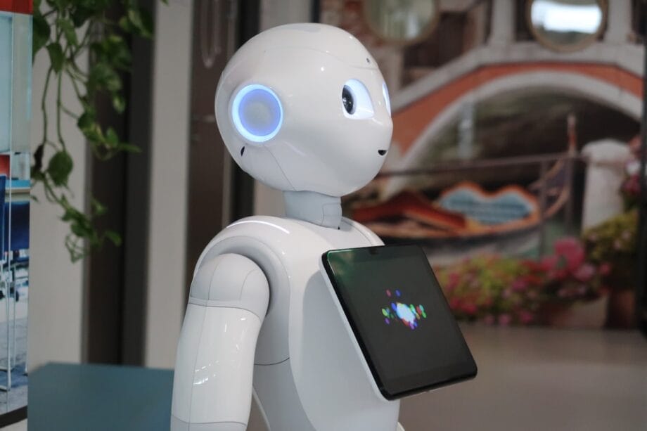 A white humanoid robot with a touchscreen display on its chest stands indoors near a colorful mural.