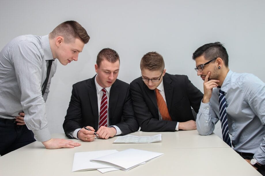 Four men in business attire review documents together at a white table in a meeting room.