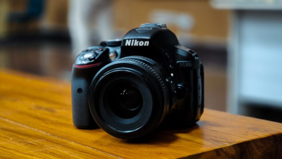 A Nikon D3400 DSLR camera with a lens is placed on a wooden surface indoors.