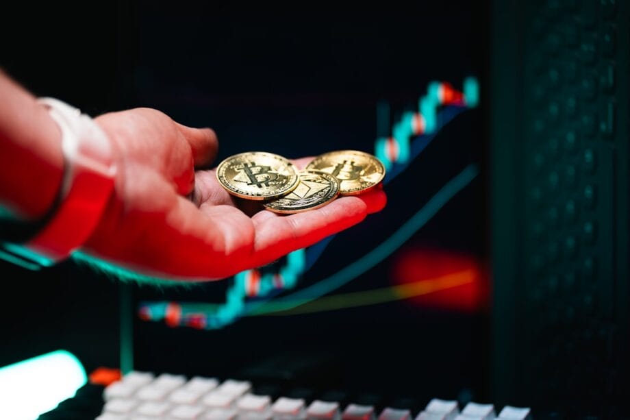 A hand holds three Bitcoin coins in front of a computer screen displaying a colorful cryptocurrency price chart.