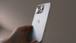 A hand holds a silver iPhone with three rear cameras against a neutral background.
