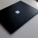 A closed black Microsoft Surface laptop sits on a round white table, partially lit by natural light.